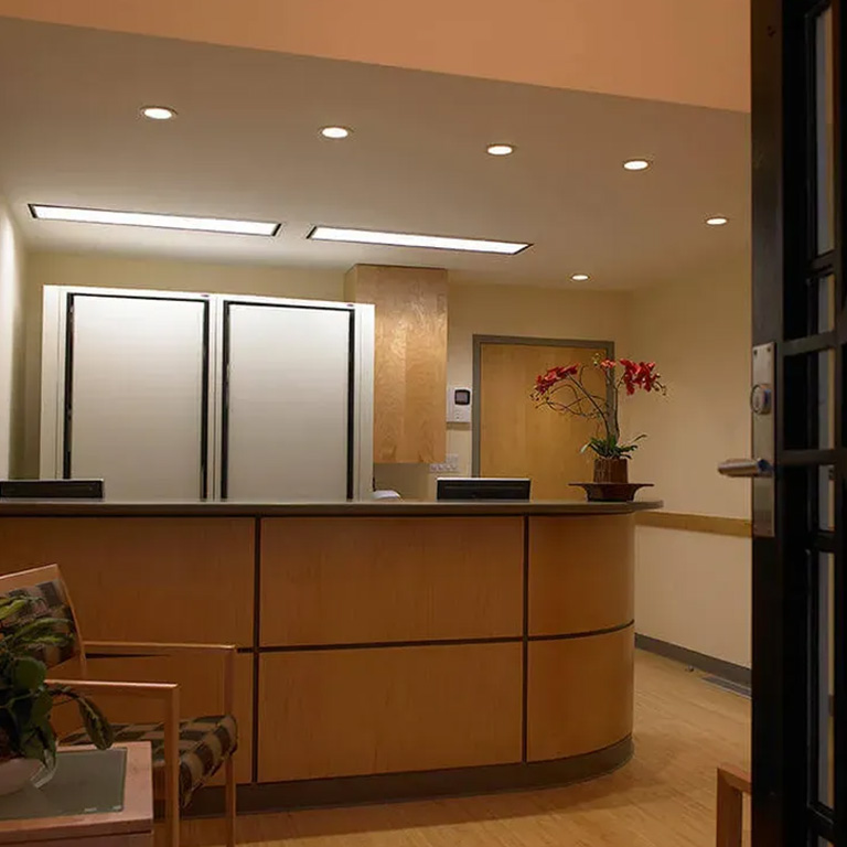 The image shows an interior space with a reception area featuring a counter, a potted plant, and a desk with a computer monitor. There is a door leading into the room and a window allowing natural light to enter.
