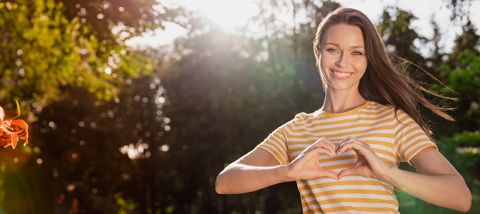 The image shows a woman making a heart gesture with her hands against a sunlit background.
