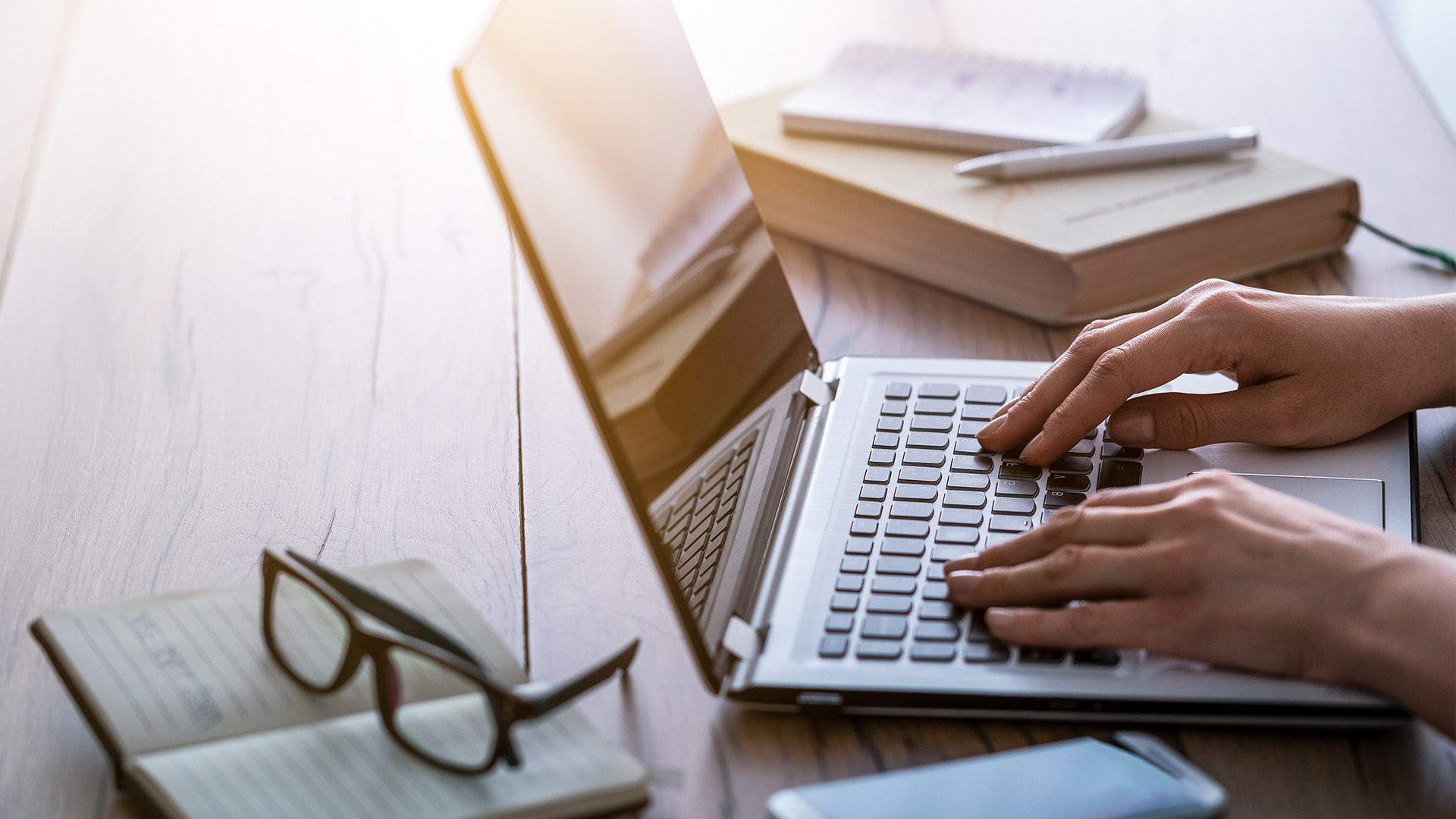 A person typing on a laptop computer at a desk with books, papers, and other office items around them.