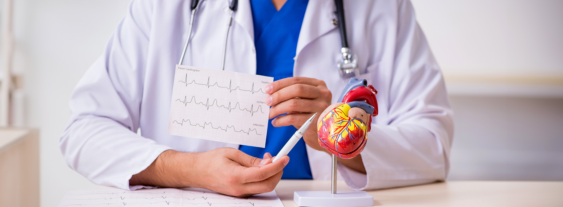 A male doctor sits at a desk with a stethoscope around his neck, holding a cardiac monitor displaying an EKG reading, wearing a white lab coat, and looking directly at the camera.