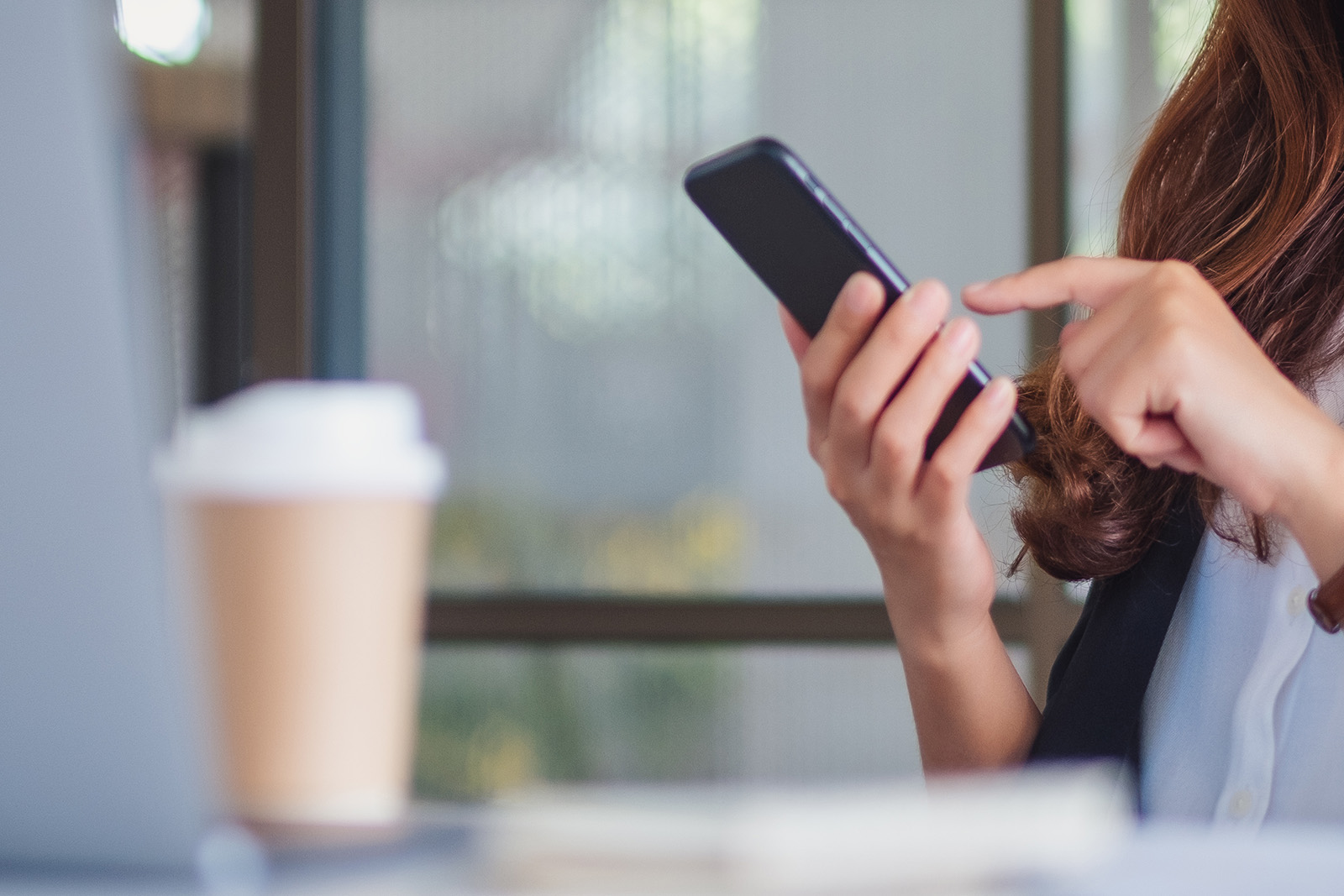 The image shows a person using a smartphone while seated at a desk with a laptop and coffee cup visible in the background, indicating a professional setting.