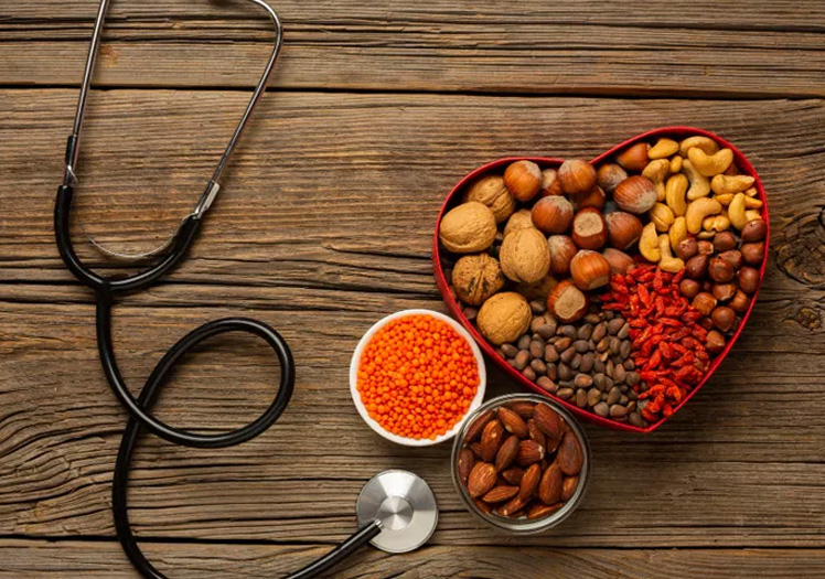 The image shows a heart-shaped container filled with various nuts and dried fruits, accompanied by a small bowl of red seeds, a stethoscope, and a bottle labeled  red peppercorns,  all arranged on a wooden table against a blurred background.