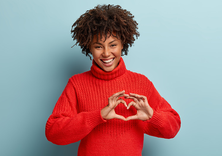 A woman with curly hair is smiling and making a heart shape with her hands against a blue background.