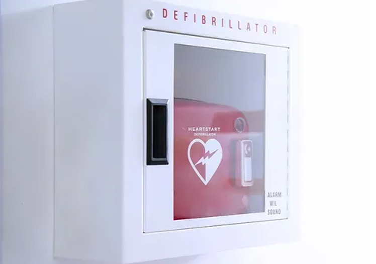 An image featuring a red emergency defibrillator with a heart symbol on its front, mounted on a white wall, next to a small cardboard box with a lock.