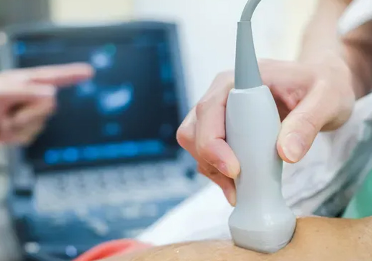 The image shows a medical setting with a person holding an ultrasound device over a patient s abdomen, where a monitor displays the ultrasound scan.
