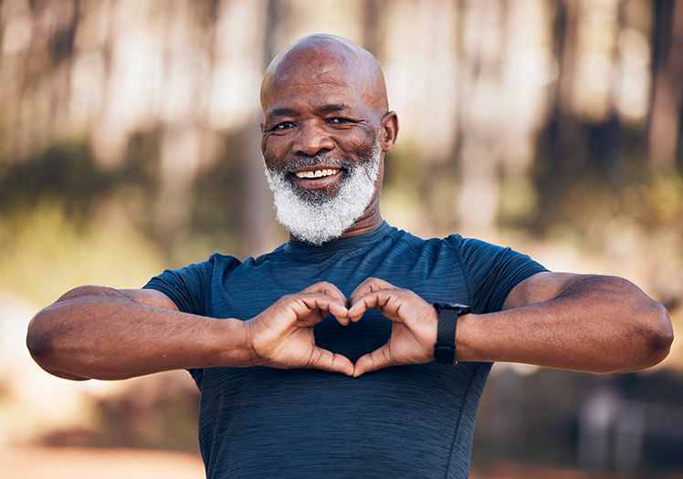 The image features a man with a beard and mustache, wearing a dark top and shorts, standing outdoors with his arms spread wide in a heart shape.