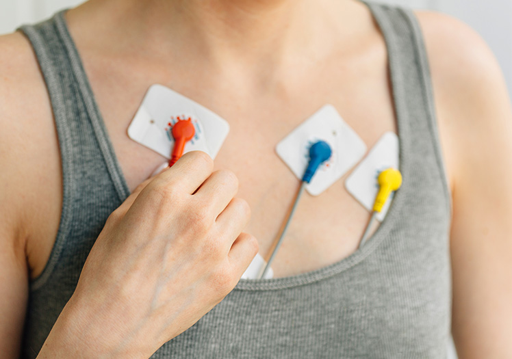 A person wearing a gray tank top holds an ECG monitor with red and yellow wires attached to their chest.