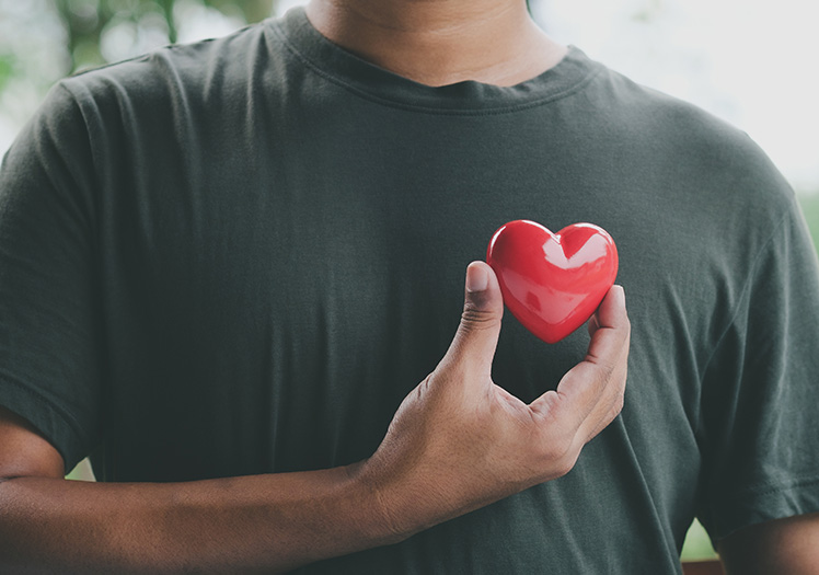 A person with their hand on their chest holding a red heart with a finger through it against a blurred background.