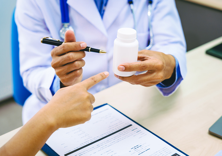 A healthcare professional, possibly a pharmacist or doctor, is holding a bottle of medication and pointing at it with a pen while seated at a desk in an office setting.
