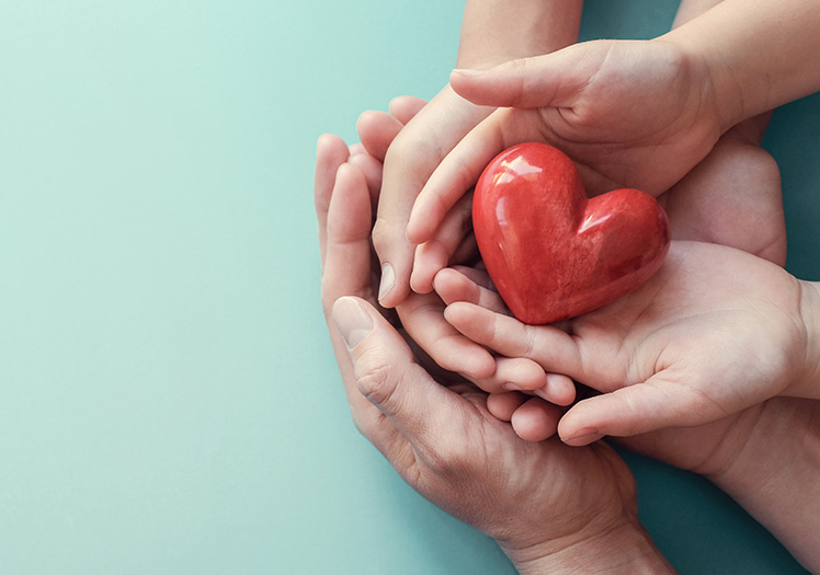 The image shows hands holding a red heart, symbolizing love and care, with fingers gently placed on top of the heart.