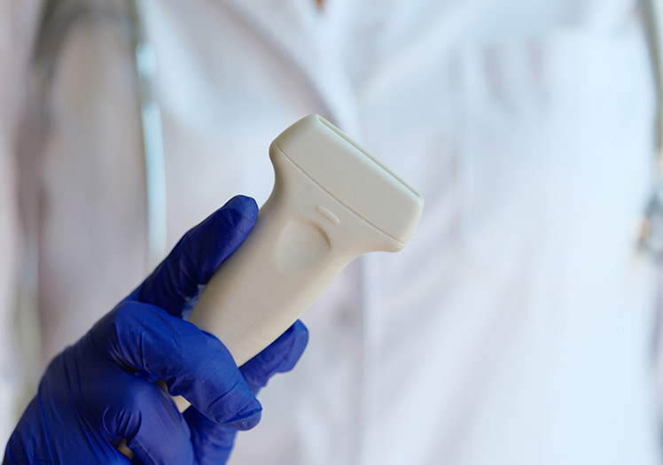 An image of a hand holding a medical device with blue gloves on, against a blurred background featuring a white lab coat.