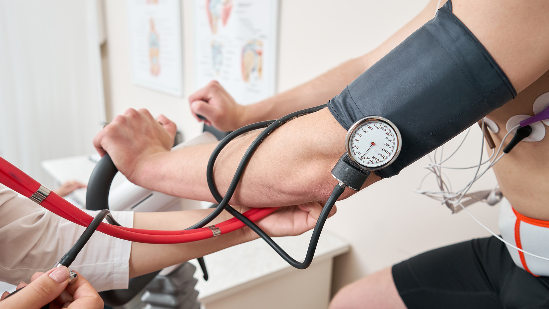 A person with a blood pressure monitor strapped to their arm, wearing a red shirt and black pants, is seated next to a large red and black blood pressure machine while another person, wearing a white coat, is standing behind them, holding a clipboard.