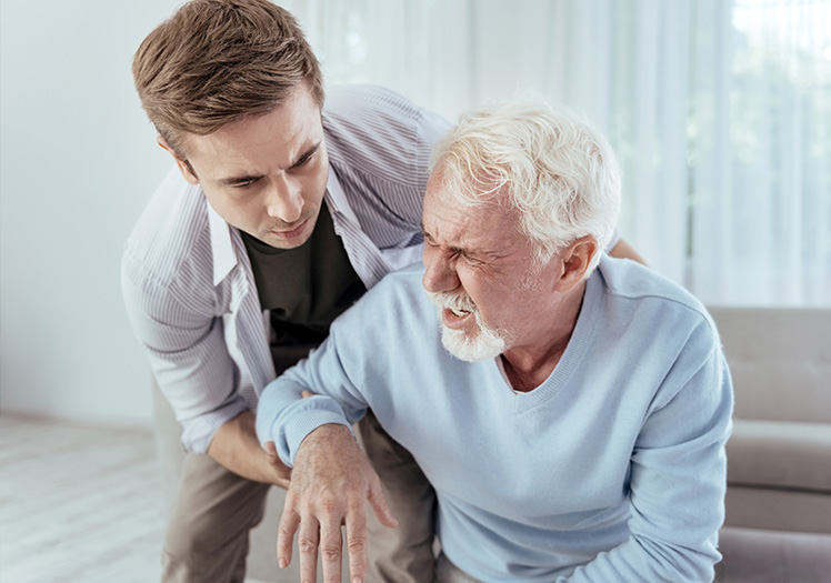 A man with white hair sitting on a couch, looking up at another man standing next to him with his hand on the older man s shoulder, who appears to be speaking or gesturing.