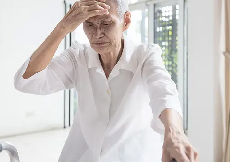 An elderly woman with her hand on her head, possibly in pain or discomfort.