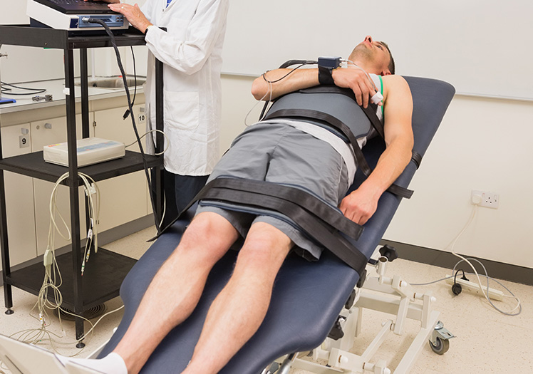 The image shows a man lying on his back on a medical examination table with his legs strapped apart, wearing a black vest, hooked up to various medical devices, while a healthcare professional stands next to him observing the equipment and the patient s condition.