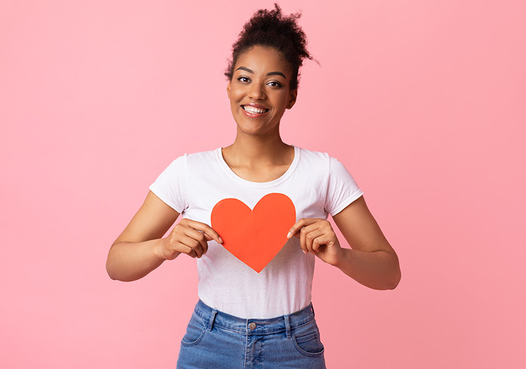 A woman holding a red heart with both hands against a pink background.